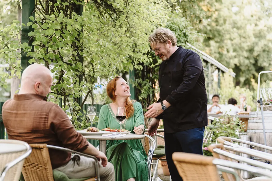 Der Koch im schwarzen Kochhemd im Gespräch mit zwei Gästen an einem Tisch auf der grün bewachsenen Terrasse des Bistro Schaarschmidt