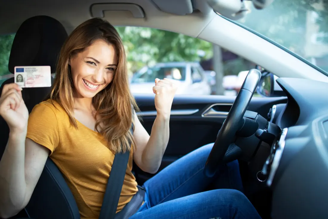 Junge Frau am Steuer eines Autos hält lachend ihren neuen Führerschein in die Kamera und ballt die Faust vor Freude